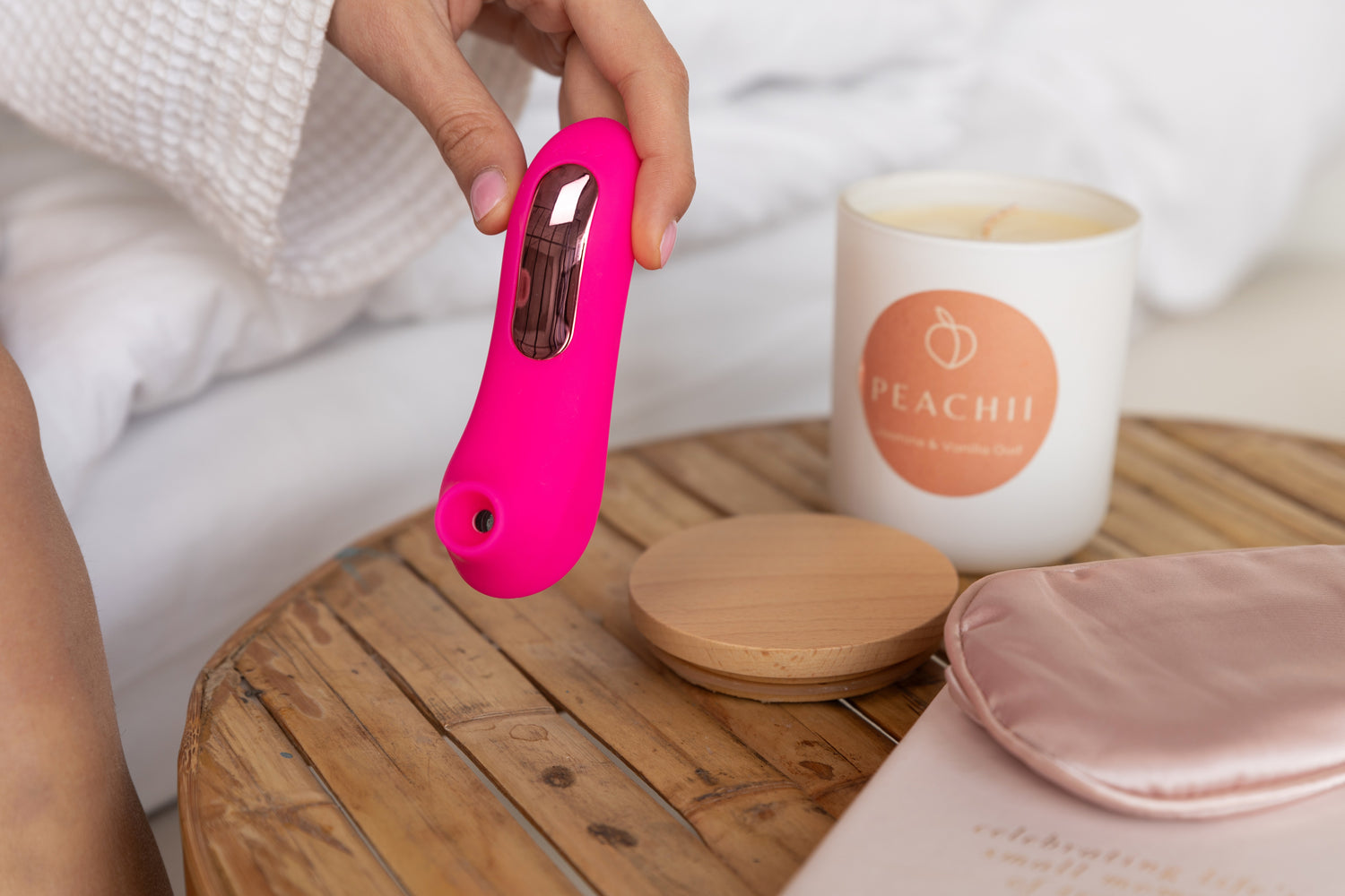 A person in a white robe holds Peachii's 'Little Pink' on a wooden table next to a Peachii scented candle. This cozy scene captures a moment of self-care and relaxation, enhanced by the soothing aroma of the candle. Peachii's commitment to sustainability shines through with their sex toy recycling program, ensuring that products like 'Little Pink' contribute to a greener planet.