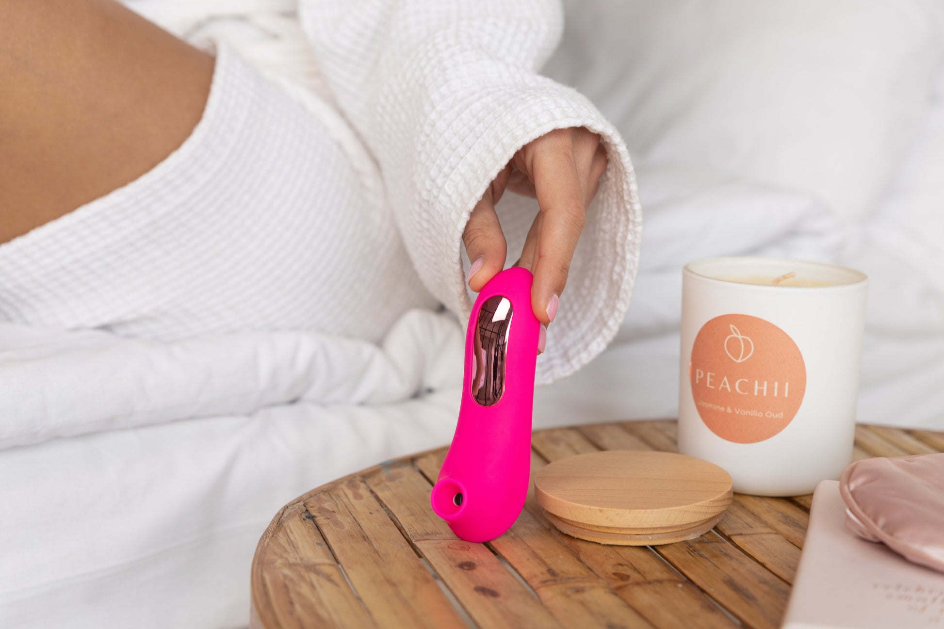 A person in a soft robe settles into a peaceful corner with a "Little Pink" device by Peachii, complemented by the warm glow of a scented candle, promising a relaxing self-care experience.