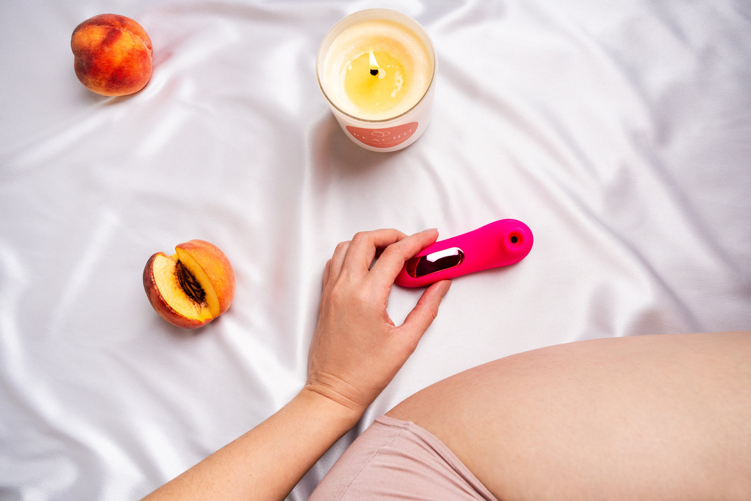 Laid out on a silky fabric, a person holds the "Little Pink" device by Peachii, flanked by lush peaches and a softly lit scented candle, weaving a narrative of sensory indulgence and self-care.