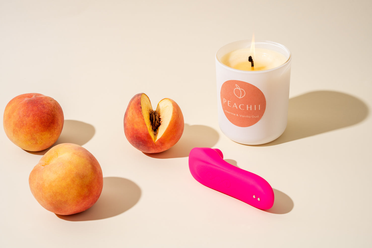 A serene setting captures the essence of self-care with Peachii's "Little Pink" device elegantly placed beside a candle scented with Jasmine & Vanilla Oud, complemented by fresh peaches. This scene evokes a relaxing afternoon devoted to sensory delight and personal wellness.