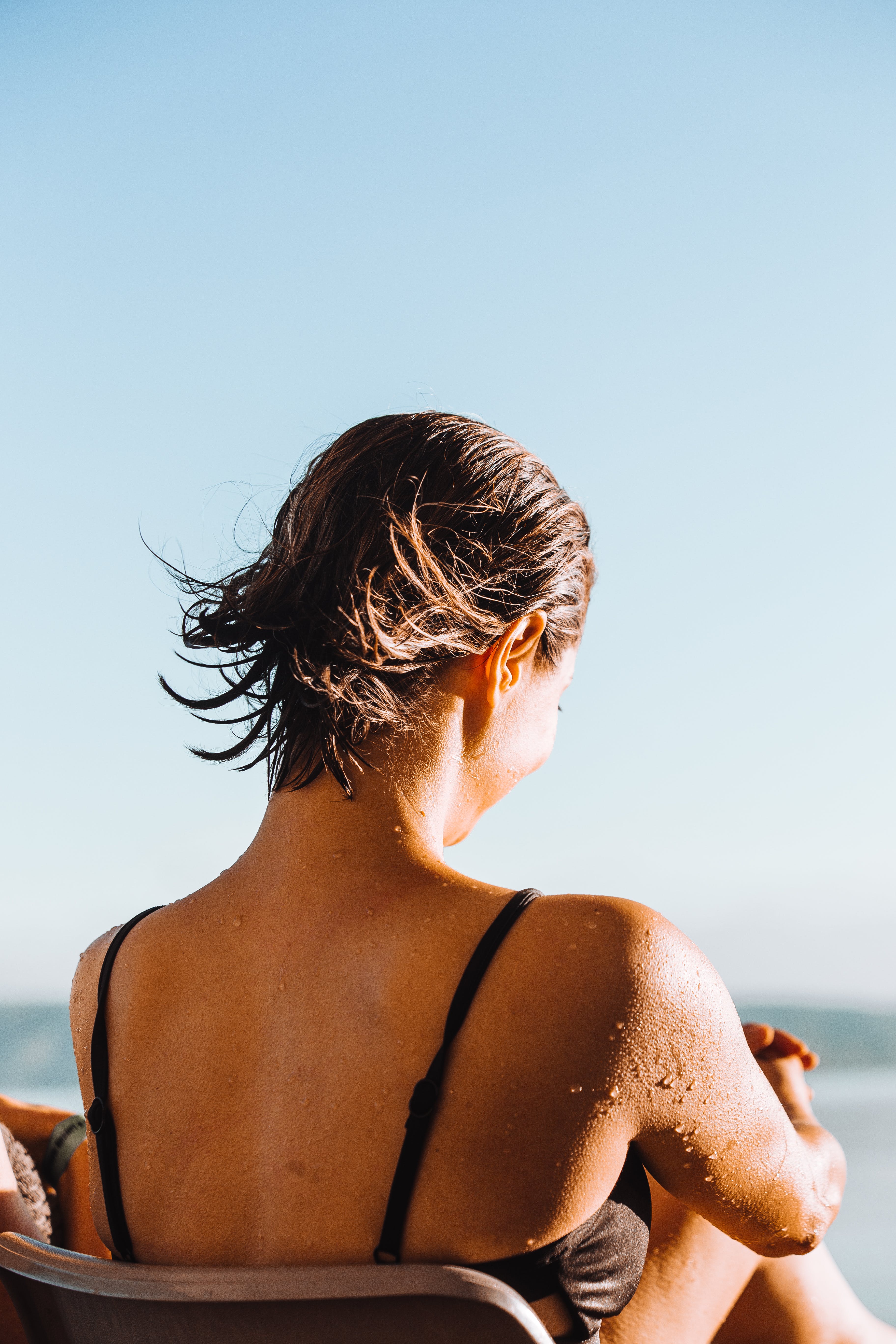 A woman relaxes by the beach, her back glistening with water droplets under the sun. She reflects on her serene moment with Peachii's 'Little Pink', a symbol of self-care and holistic wellness. This peaceful scene highlights Peachii's commitment to sustainability through their sex toy recycling program, ensuring products like 'Little Pink' contribute to a healthier planet.